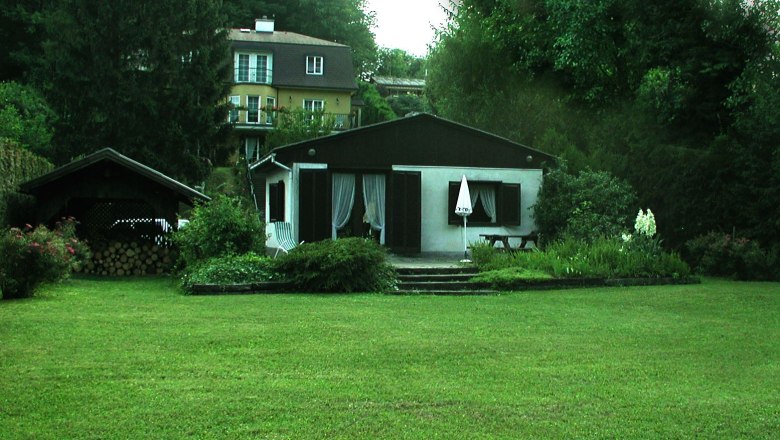 Ein kleines Haus mit Garten und Terrasse, umgeben von B&auml;umen und einem gr&ouml;&szlig;eren Geb&auml;ude im Hintergrund.