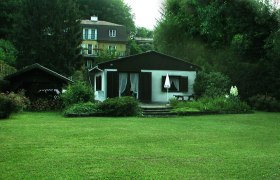 Ein kleines Haus mit Garten und Terrasse, umgeben von B&auml;umen und einem gr&ouml;&szlig;eren Geb&auml;ude im Hintergrund.