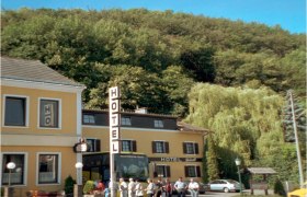 Ein gelbes Hotelgeb&auml;ude mit einem gro&szlig;en Hotelschild und Menschen davor, umgeben von B&auml;umen.