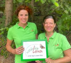 Irene Obetzhofer und Doris Lackner in Wald mit Natürlich Lernen Schild