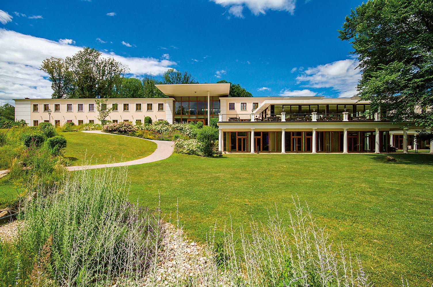 A modern building with large windows and a terrace, surrounded by a well-tended garden and trees under a blue sky.